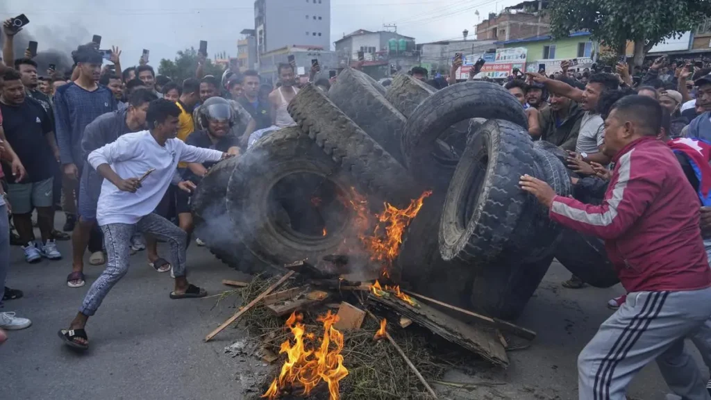 A large crowd of young people, often referred to as "Gen Z," is gathered on a street in Nepal to protest. Many are holding placards with slogans and waving the red and blue national flags of Nepal. The demonstration is a protest against the government's ban on social media and wider issues of corruption and nepotism.