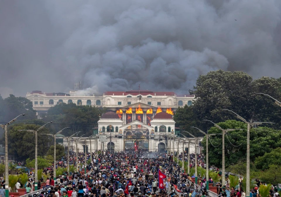 A massive crowd of protestors fills the street leading up to a large, white building with a classical facade. The lower part of the building is engulfed in bright orange flames and heavy smoke pours from the roof and upper windows. The sky is dark with a thick plume of black smoke rising from the building, obscuring the sky. The foreground is crowded with people, some holding signs, others on motorbikes, all facing the burning building. The street is lined with green trees and streetlights on either side.